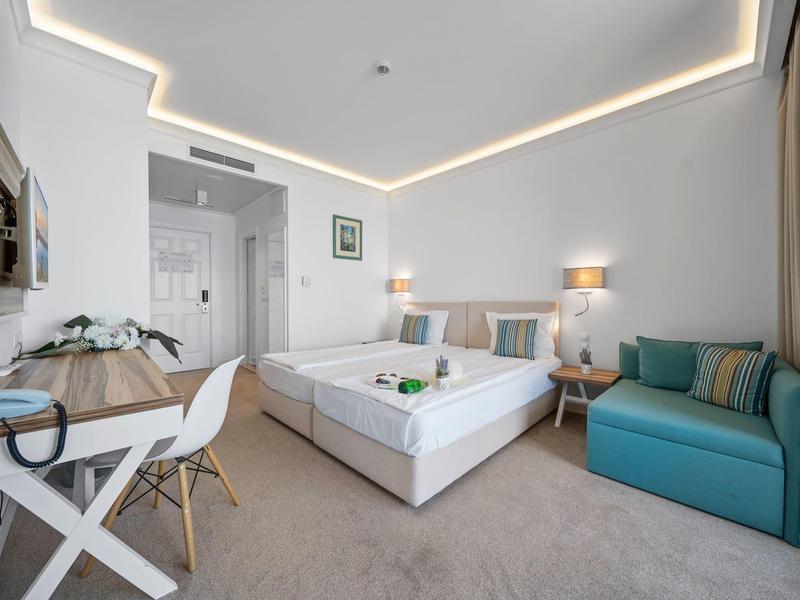 Moderní hotelový pokoj s velkou postelí, psacím stolem, zeleným křeslem a nepřímým osvětlením.