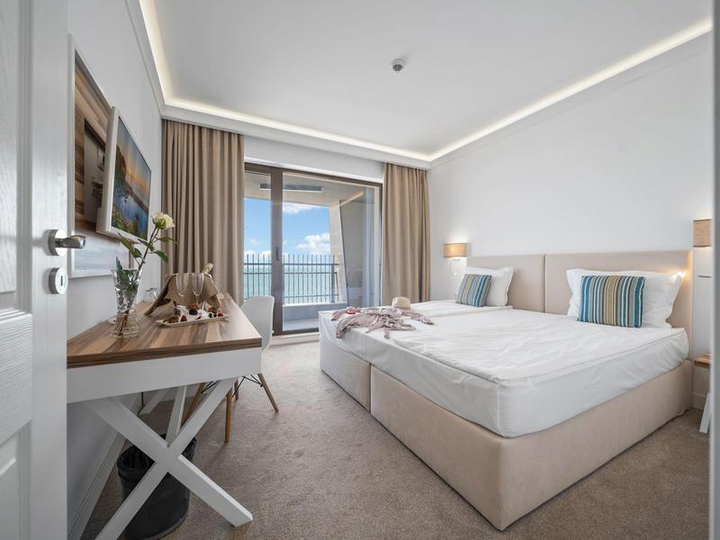 Moderní hotelový pokoj s manželskou postelí, stolem a výhledem na moře přes velká okna.