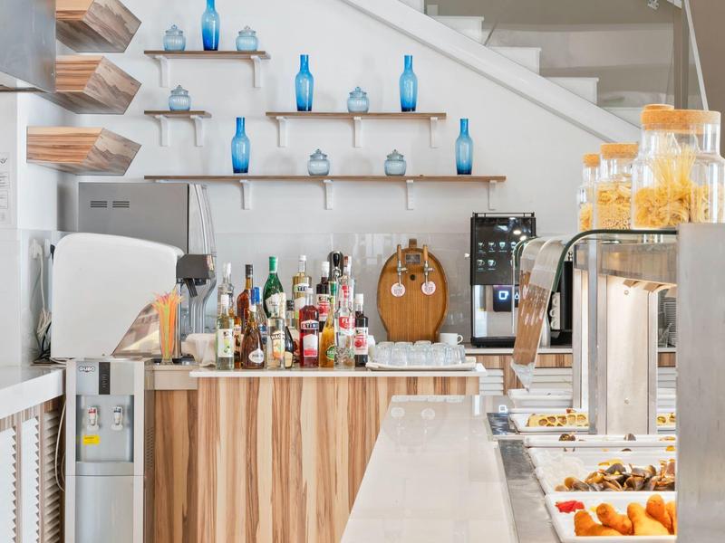 Moderní hotelový bar s dřevěným pultem, nápoji, zásobníkem na džus a modrými vázami na policích pod schodištěm.