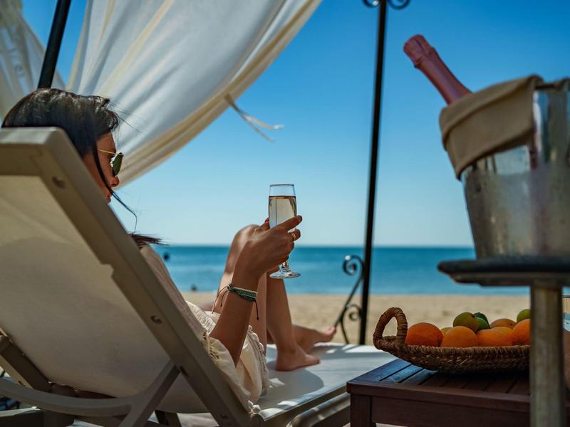 Osoba odpočívající na lehátku na pláži s nápojem v ruce a výhledem na moře v pozadí.