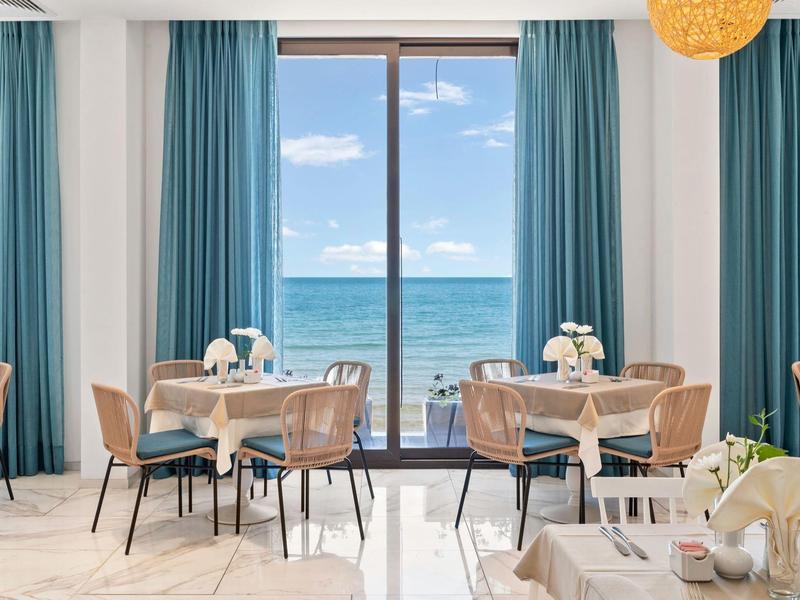 Světlá jídelna s modrými závěsy a výhledem na moře skrz velká okna.