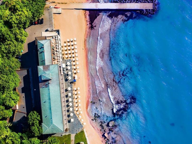 Letecký pohled na pláž s hotelem, lehátky a čistou modrou vodou vedle husté zeleně.