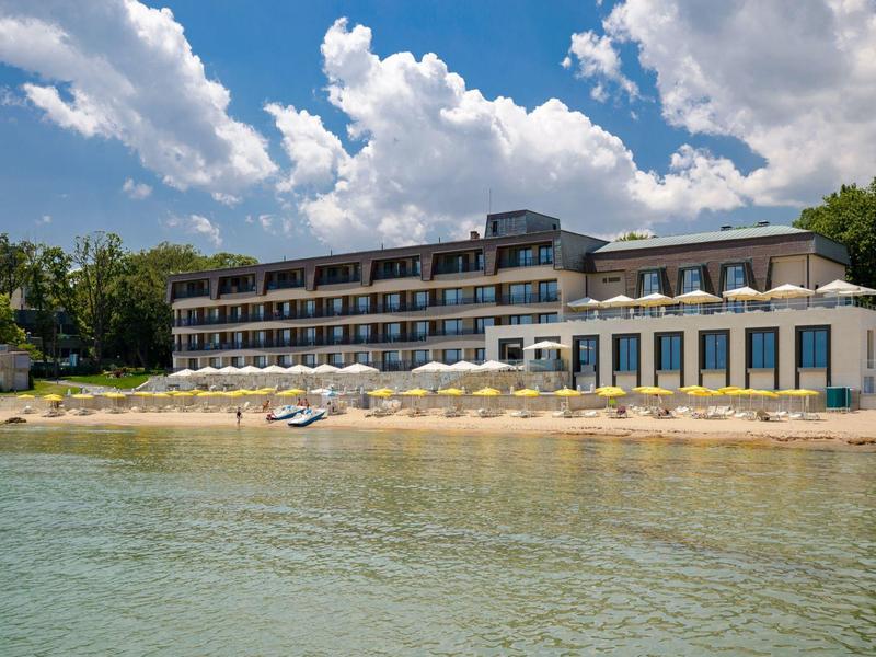 Moderní hotel na pláži s lehátky a modrou oblohou s mraky.