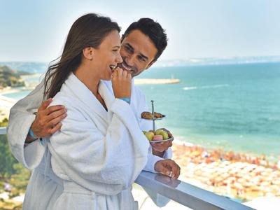 Pareja con batas disfrutando desayuno en balcón con vista al mar y playa.