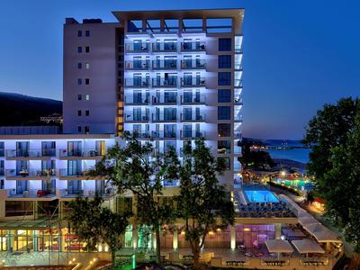 Edificio de hotel iluminado con habitaciones con balcón y área de piscina al anochecer.