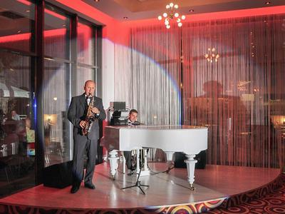 Musicians play saxophone and piano on a stage in a hotel with red lighting.