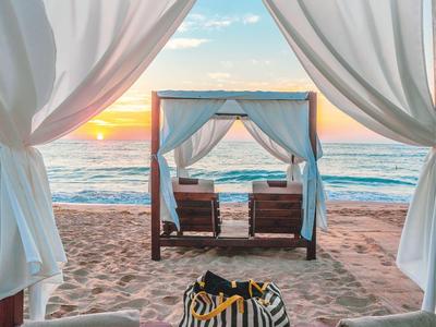 Cama de playa con cortinas blancas y atardecer sobre el mar al fondo.