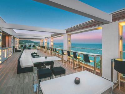 Moderne Dachterrasse mit Sitzbereichen, Barhockern und Blick auf den Sonnenuntergang über dem Meer.