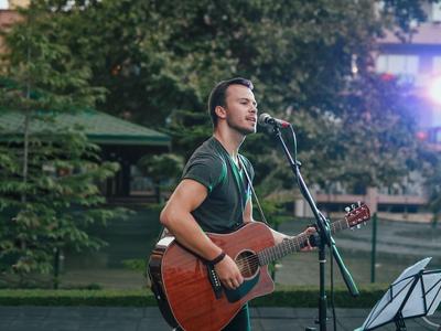Man playing guitar and singing outdoors with microphone, trees, and lights in background