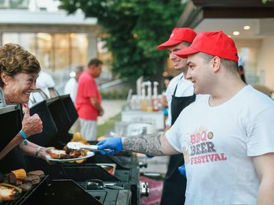 Two men wearing red caps serve food to a woman at an outdoor barbecue stand.