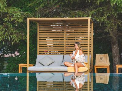 Mujer sentada en un lounge exterior junto a la piscina con árboles al fondo.