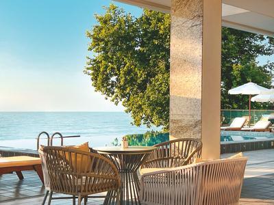 Terrace with chairs and table overlooking the pool and sea under a clear sky.
