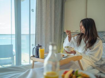 Mujer en bata desayunando en habitación de hotel con balcón y vista al mar.