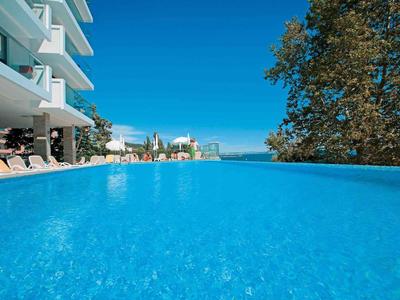 Niebieski basen hotelowy z leżakami i widokiem na drzewa oraz czyste niebo.