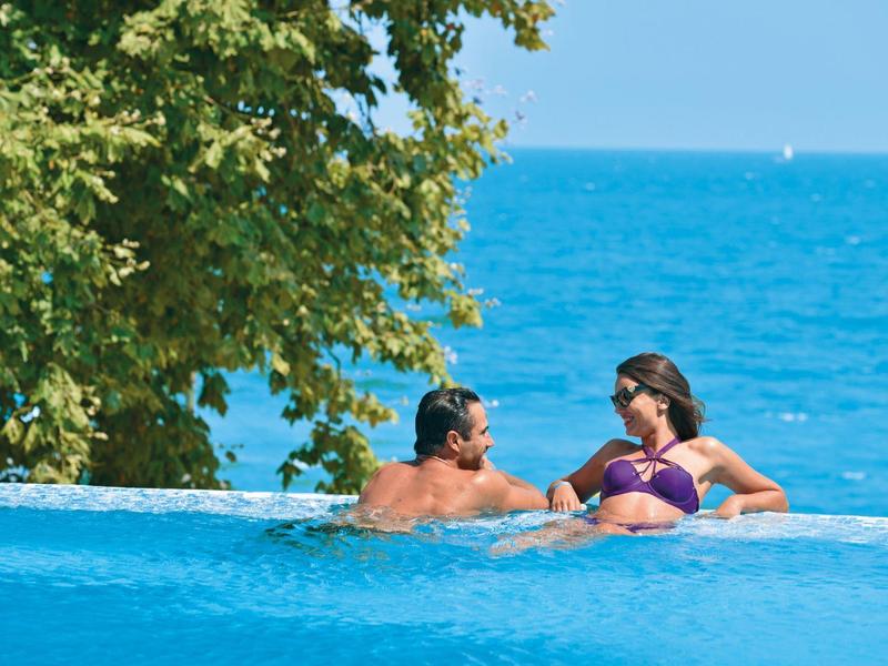 Pareja relajándose en una piscina infinita con vista al mar y un árbol cercano.