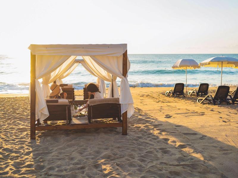 Playa con tumbonas y una cama con dosel al atardecer junto al mar.