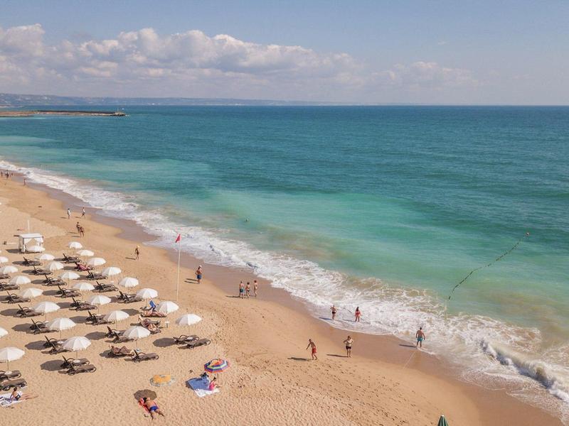 Playa de arena con filas de sombrillas y personas descansando en el mar y la orilla.