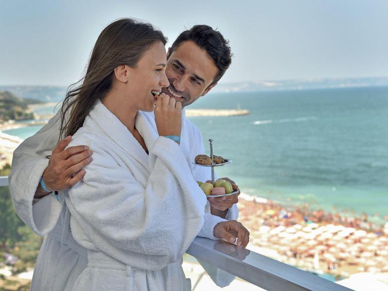 Pareja en batas disfruta desayuno con vista al mar en un balcón.