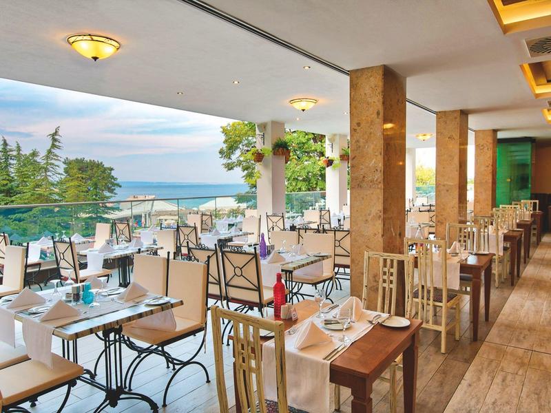 Terraza de restaurante con mesas y sillas, vista al mar y bosque, ambiente iluminado y acogedor.