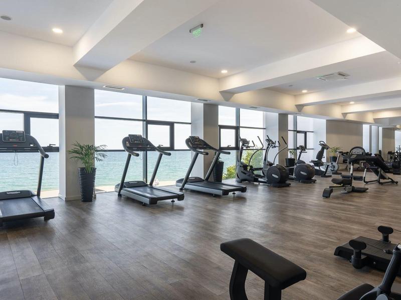 Gimnasio luminoso con cintas de correr y bicicletas, grandes ventanas y suelo de madera.