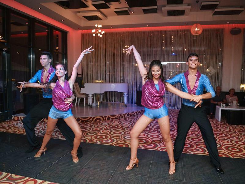 Cuatro bailarines con trajes brillantes realizan una presentación de baile en pareja en un salón de hotel.