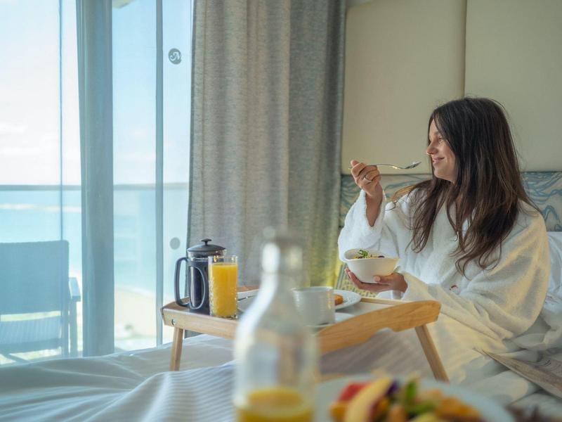 Mujer en bata desayunando en habitación de hotel con balcón y vista al mar.