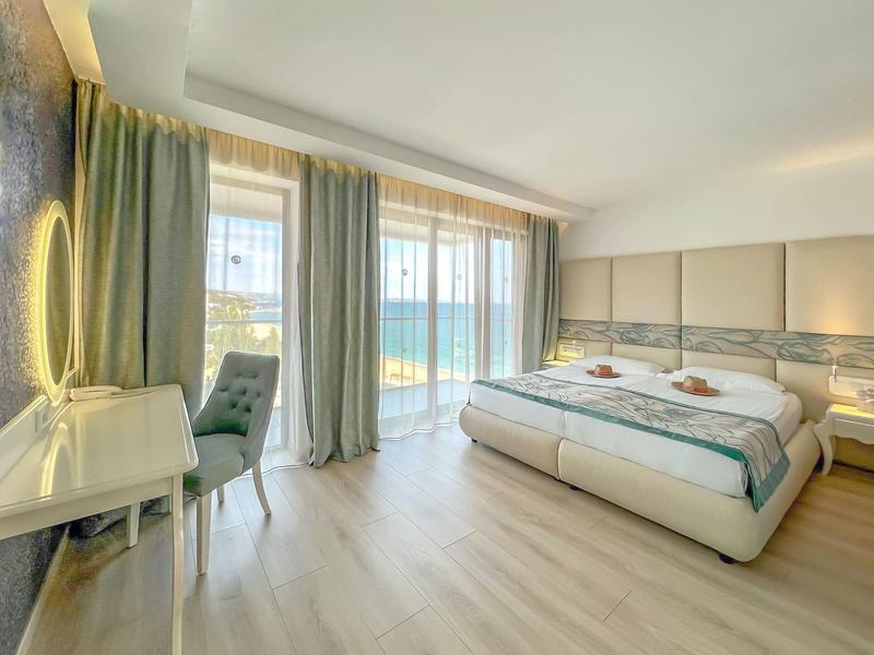 Habitación de hotel luminosa con cama doble, escritorio y vista al mar a través de grandes ventanas.