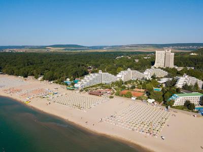 Vue aérienne d'une plage de sable avec des hôtels et un arrière-plan boisé sous un ciel clair.