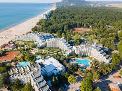 Vue aérienne d'une station avec plusieurs bâtiments, piscines et une longue plage de sable à côté de la forêt.