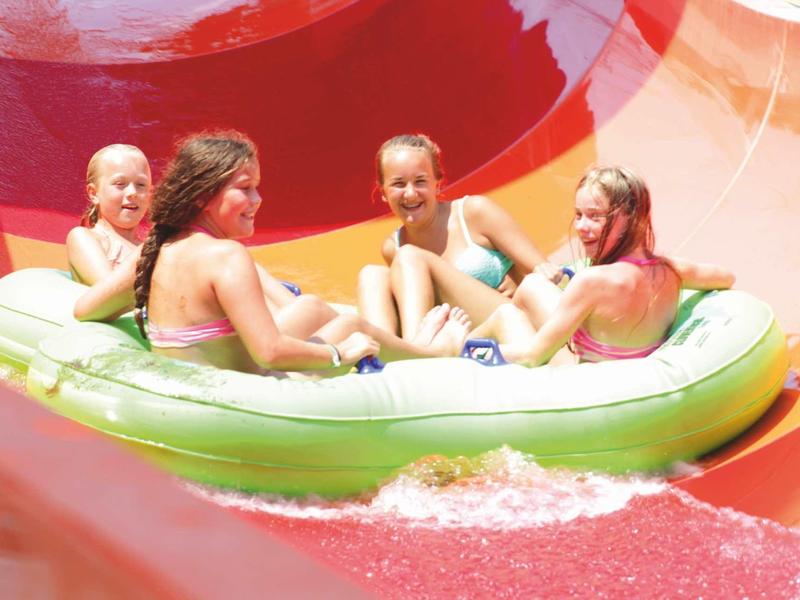 Four children slide together down a water slide on an inflatable raft.
