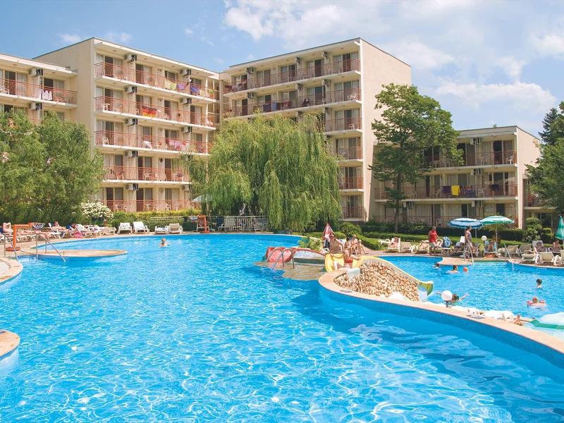 Hotel complex with large outdoor pool, sun loungers, and several multi-story buildings on a sunny day.