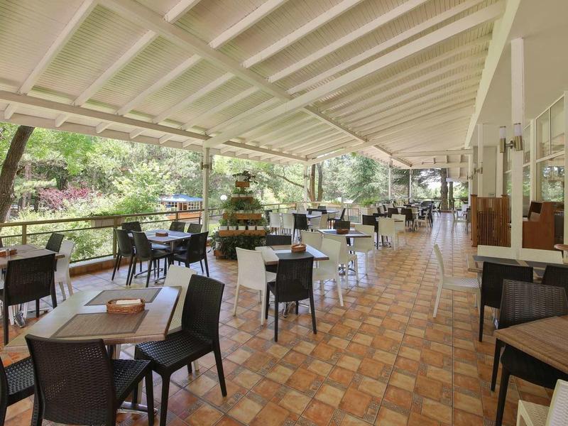 Covered outdoor restaurant terrace with tables and chairs, surrounded by trees and plants.
