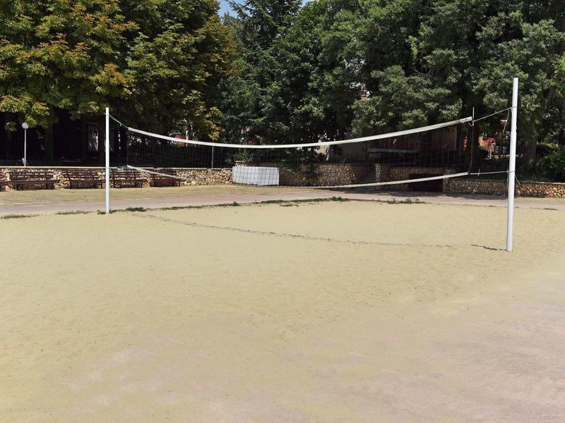 An empty sand volleyball court surrounded by trees and a wall in the background.