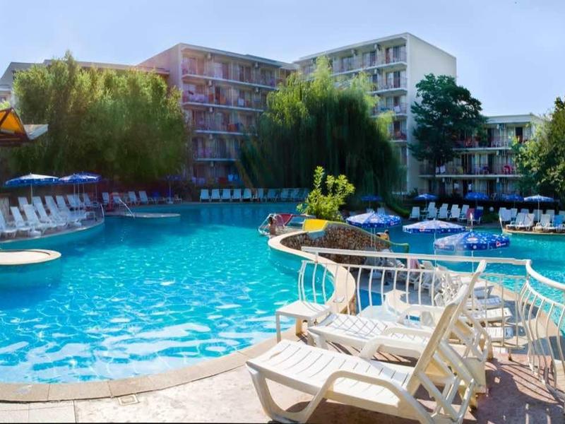 A large outdoor swimming pool with sun loungers and umbrellas in front of a hotel and trees.