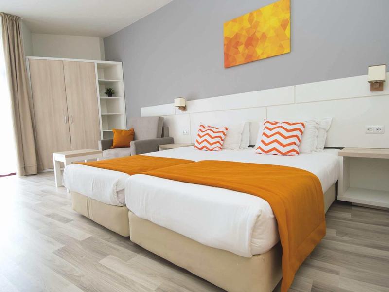 Modern hotel room with two single beds, orange bed runners, and a wardrobe.