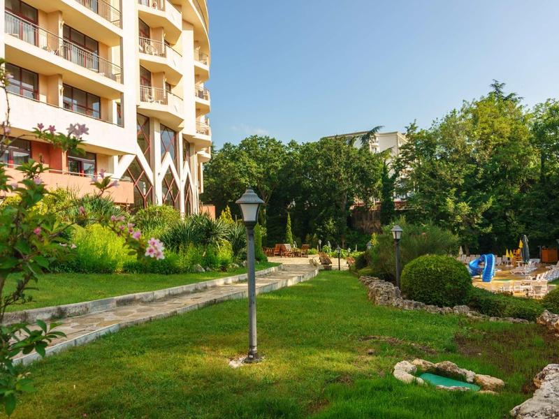 Green hotel garden with pathways, seating areas, and flower decorations under clear sky.