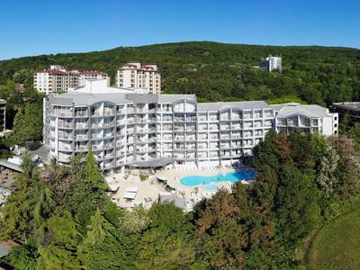 Grand hôtel blanc avec piscine, entouré d'arbres et de collines vertes en arrière-plan.