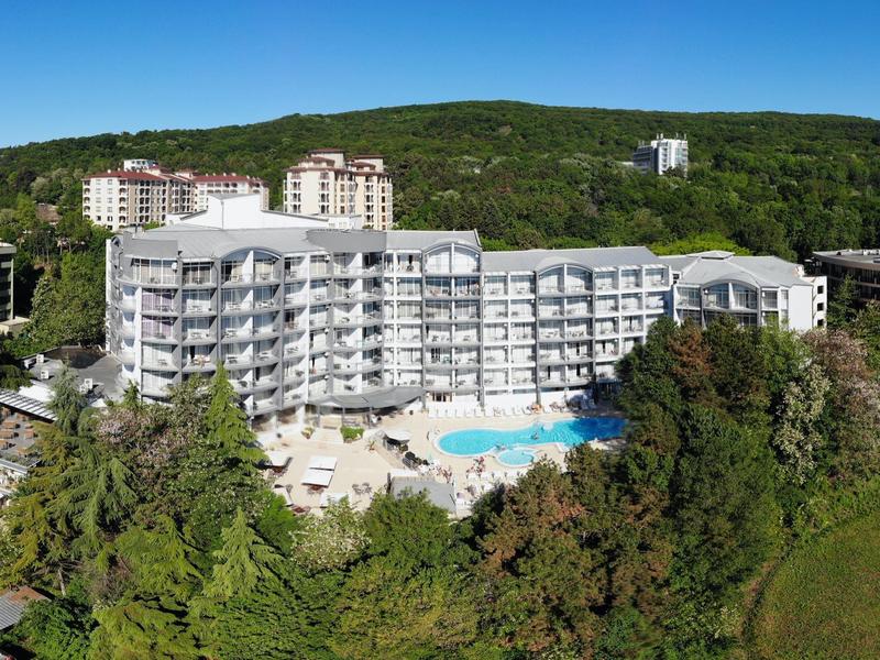 Grand hôtel blanc avec piscine, entouré d'arbres et de collines vertes en arrière-plan.