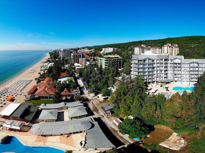 Vue panoramique d'un hôtel en bord de mer avec piscine et collines verdoyantes en arrière-plan.