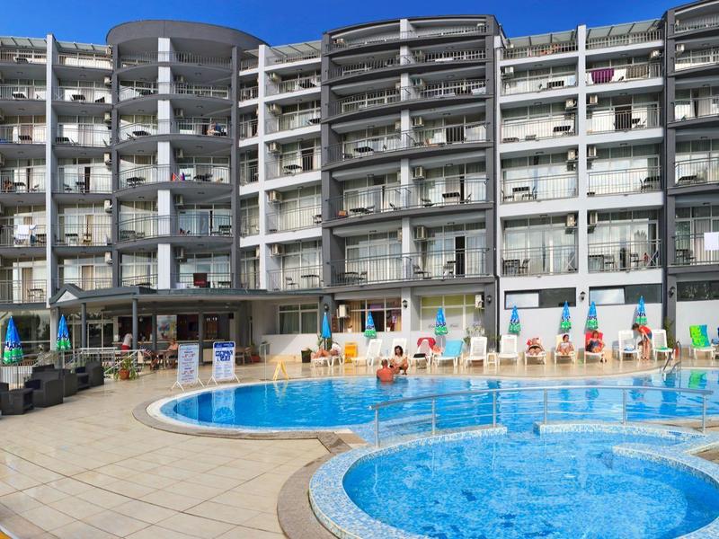Grand hôtel à plusieurs étages avec une piscine ronde au premier plan sous un ciel bleu clair.