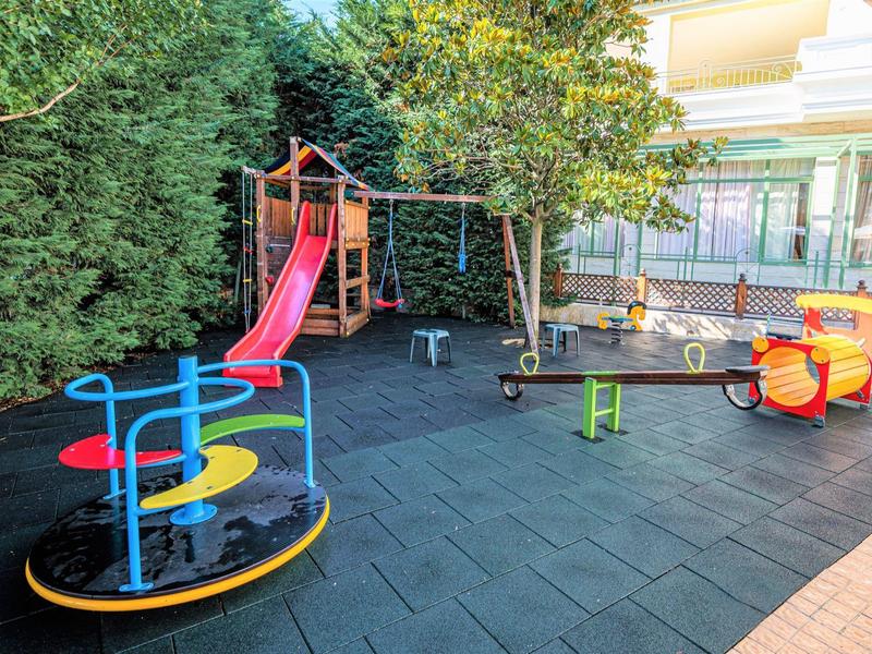 Area giochi colorata per bambini con scivolo, giostra e altalena su tappetini di gomma accanto a un hotel