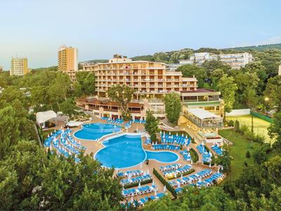 Groot hotel met meerdere zwembaden en veel ligstoelen omgeven door groen.