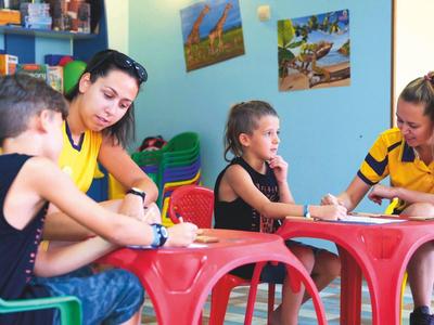 Kinderen zitten aan kleine tafels en kleuren met volwassenen in een kleurrijke kamer.