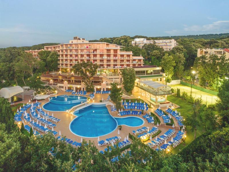 Groot hotel met meerdere zwembaden en ligstoelen, omgeven door weelderig groen.