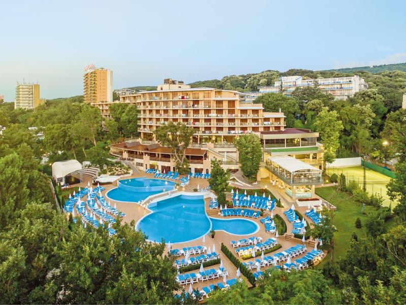 Groot hotel met meerdere zwembaden en veel ligstoelen omgeven door groen.