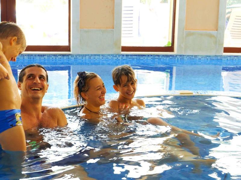 Gezin met twee kinderen ontspant in binnenzwembad bij daglicht naast grote ramen.