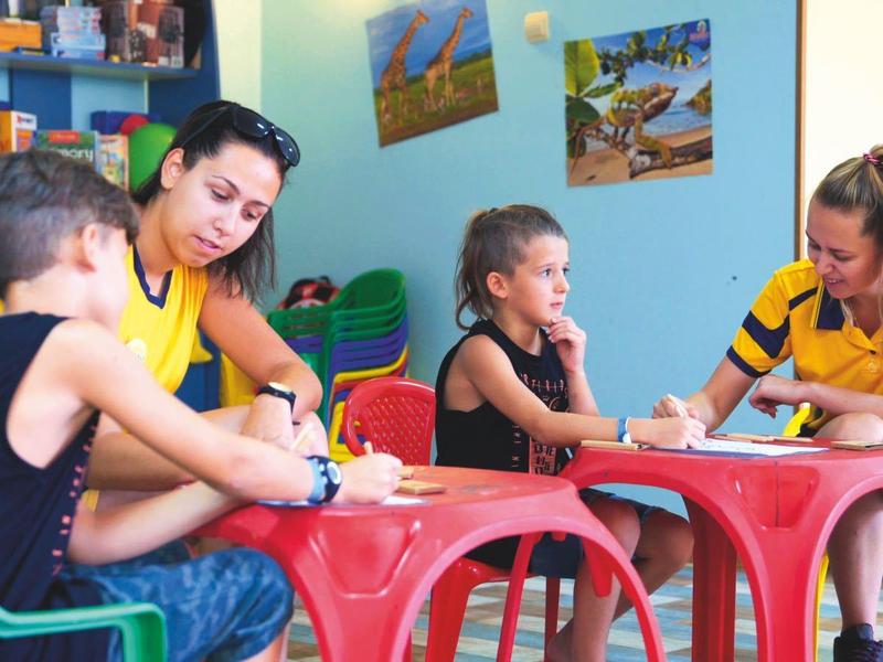 Kinderen zitten aan kleine tafels en kleuren met volwassenen in een kleurrijke kamer.