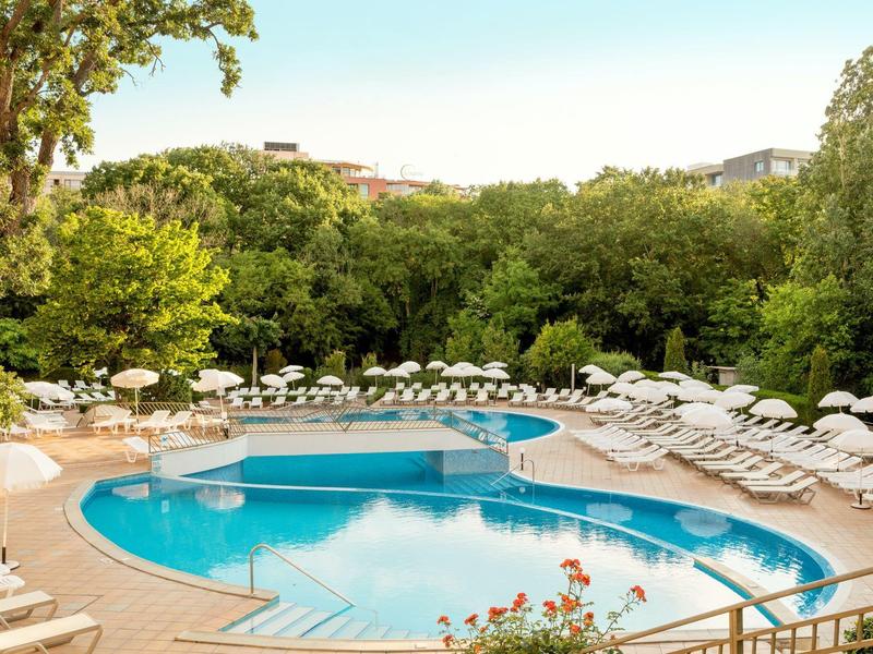 Rond buitenzwembad met witte ligstoelen en parasols omringd door groene bomen.