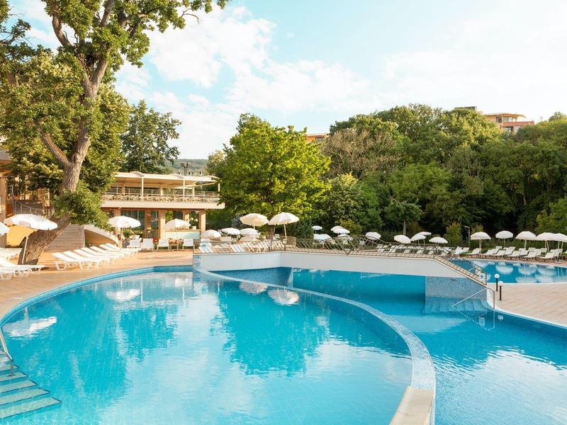 Groot rond hotelzwembad met parasols, omgeven door bomen onder een blauwe lucht.