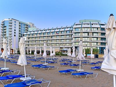 Strand mit blauen Liegen und weißen Sonnenschirmen vor einem modernen, mehrstöckigen Hotelgebäude.